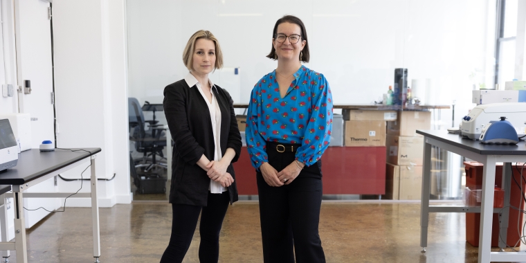 Two women, Cecilia Kushner and Beck Brachman, stand side by side in a modern, bright laboratory. One wears a black suit and heels, the other a blue blouse with a red pattern and black pants. Lab equipment and workstations are visible in the background.