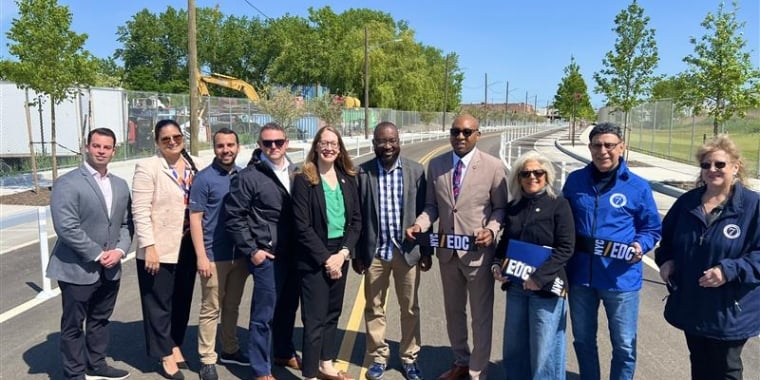 Group of ten people stand together on a newly paved road, smiling at the camera. Some are holding a ribbon with the NYCEDC logo on it. Trees and construction equipment are visible in the background under a clear blue sky.