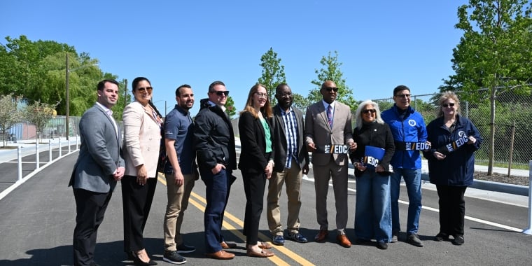Group of ten people stand together on a newly paved 132nd Street extension in College Point, smiling at the camera. Some are holding a ribbon with the NYCEDC logo on it. Trees and construction equipment are visible in the background under a clear blue sky.