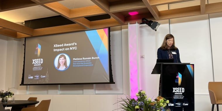 A woman stands at a podium speaking at the XSeed Award event. A large screen behind her displays the title XSeed Award’s Impact on NYC and her name, Melissa Román Burch. Flowers are arranged near the podium.