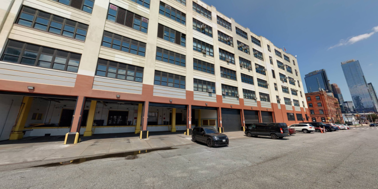 A wide, five-story industrial building with large windows and multiple garage doors at street level. Several cars are parked along the building, and city skyscrapers are visible in the background under a clear sky.