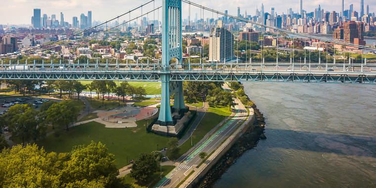 Aerial view of the Robert F. Kennedy Bridge (formerly Triborough Bridge) spanning over water, with Manhattan’s skyline in the background and green parks and roads visible below.