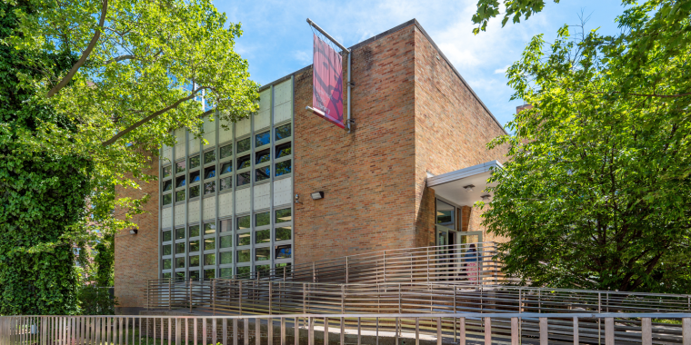 A modern brick building with large grid-like windows, a sidewalk with a metal fence in front, and leafy green trees surrounding the entrance. A red New York Public Library banner hangs near the entrance on a sunny day.