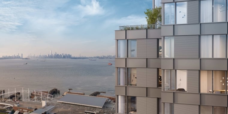 A modern high-rise building with large windows, overlooks a waterfront, with boats on the water and a New York City skyline visible in the distance under a blue sky.