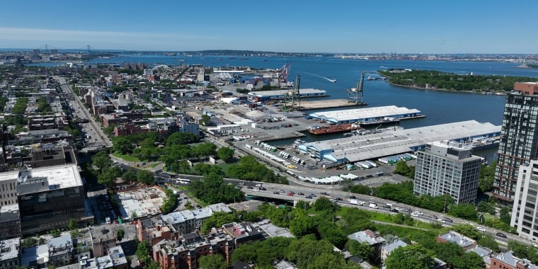 Aerial view of the Brooklyn Marine Terminal with industrial buildings, shipping docks, highways, green spaces, and a river; bridges and islands are visible in the distance under a clear blue sky.