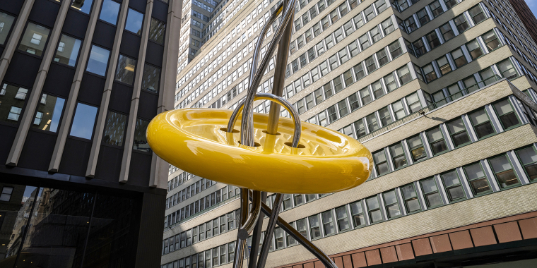 A large yellow button sculpture with silver needle and thread stands in front of tall office buildings in a city, viewed from a low angle.