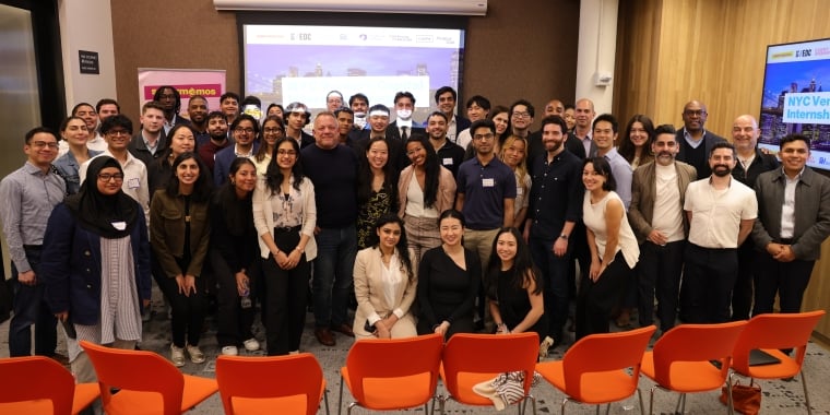 NYC Venture Capital Internship program 2025 cohort pose and smile together in a conference room with orange chairs, a projector screen, and banners in the background.