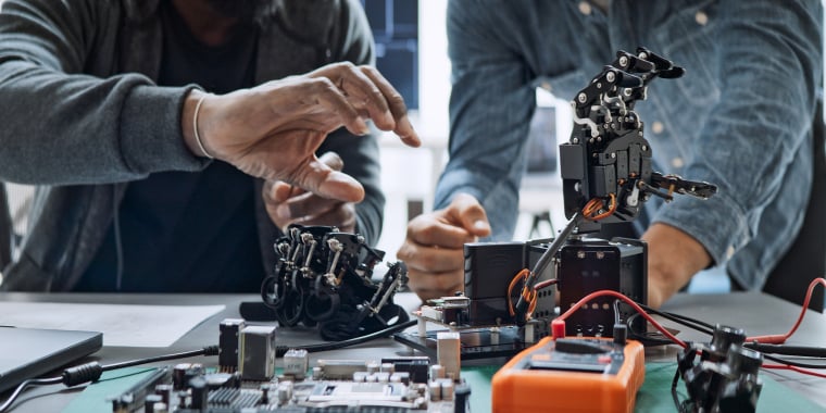 Two people work together on assembling and adjusting a robotic hand at a desk covered with electronic components, wires, and circuit boards.
