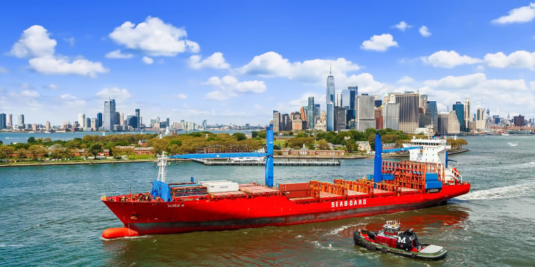 A large red cargo ship labeled Seaboard sails through the water near Manhattan, New York City, accompanied by a tugboat, with skyscrapers and blue sky with clouds in the background.