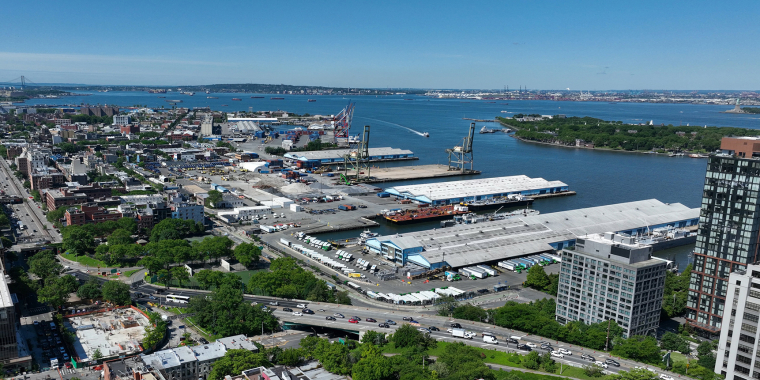 Aerial view of the Brooklyn Marine Terminal with industrial buildings, shipping docks, highways, green spaces, and a river; bridges and islands are visible in the distance under a clear blue sky.