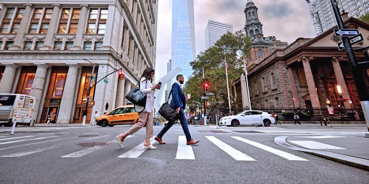 Two people cross a city street at a crosswalk, surrounded by tall buildings. The One World Trade Center tower is visible in the background. Cars, a taxi, and street signs are also present.