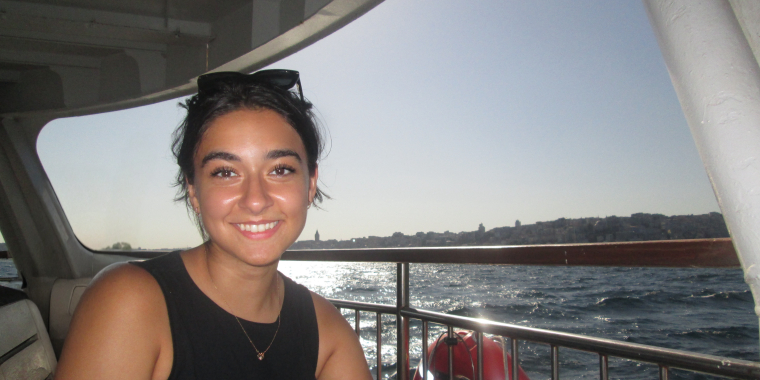 Woman with dark hair and a black sleeveless top, Leyla Arcasoy, smiles while sitting on a boat. The sun is shining brightly over the water in the background, and a cityscape is visible on the horizon.