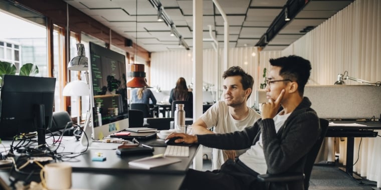 Two men sit at a desk in a modern office, collaborating as they look at a computer screen filled with code. Papers, notebooks, and a water bottle are on the desk, and other people work in the background.