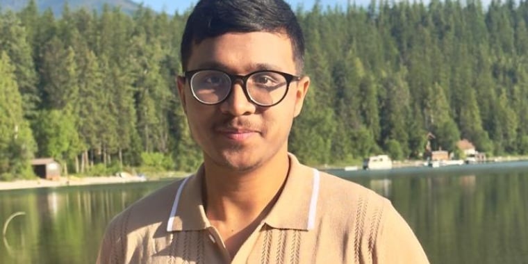 Debashrestha Nandi wearing glasses and a beige shirt stands by a lake with a forest and mountains in the background on a sunny day.
