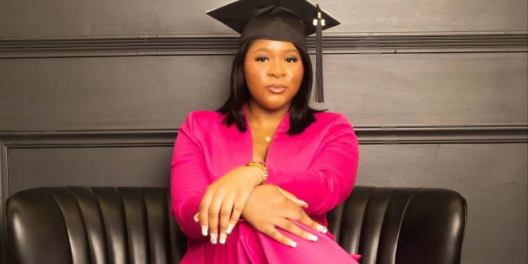 Femi Jordan wearing a bright pink suit and graduation cap, sits confidently on a black leather couch against a dark wall, looking directly at the camera with her hands crossed on her knee.