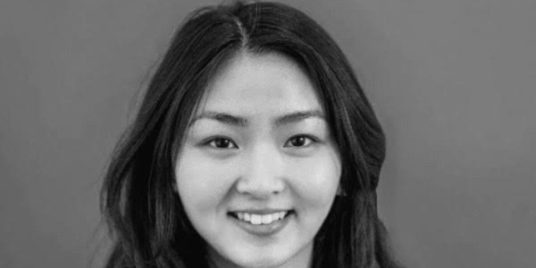 Woman with long dark hair, Justine Cheng, smiles at the camera, wearing a light-colored blazer and necklace, against a plain background. The image is in black and white.