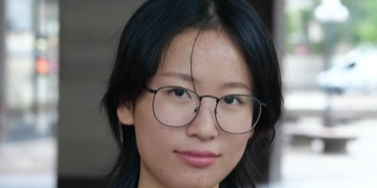 Kelly Lin with straight black hair, wearing round glasses and a dark top, looks at the camera with a slight smile. The background is blurred, showing an outdoor urban setting.