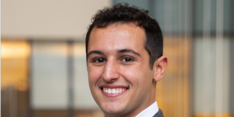 Mishah Aryeh with short, curly dark hair smiles while wearing a grey plaid suit jacket, white shirt, and pocket square. He stands indoors with a blurred office background.