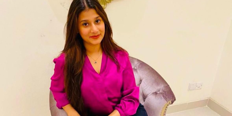 A woman with long brown hair, Samrin Alam, wearing a bright pink blouse and dark pants, sits on a light purple tufted chair in a room with cream-colored walls and a decorative mirror behind her.