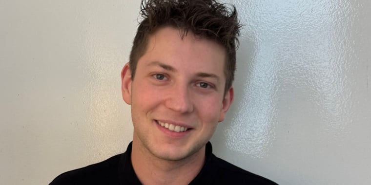 Alexandru Daniel Soroiu with short, curly brown hair and a friendly smile stands against a plain light-colored wall, wearing a black shirt.