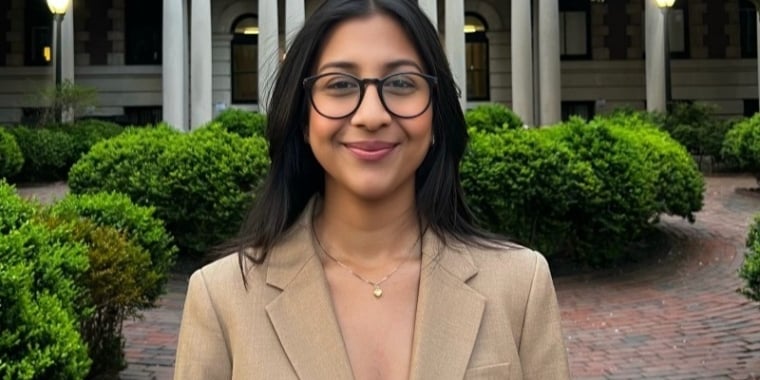 Anushka Khetawwat with long dark hair and glasses, wearing a beige blazer and a necklace, stands smiling in front of a building with columns and green bushes.