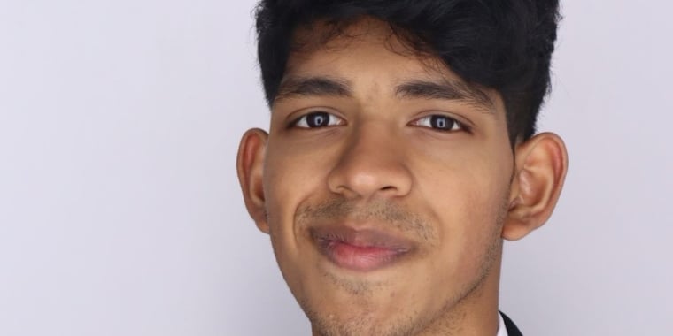 Dev Iyer with short dark hair, wearing a black blazer and a white shirt, smiles slightly while looking at the camera against a plain light background.