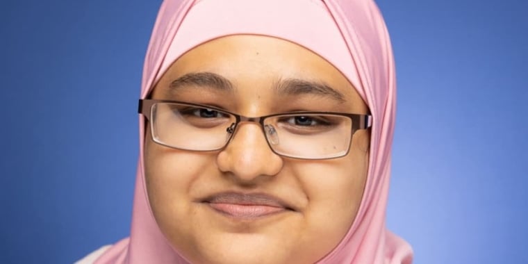 Samira Chowdhury wearing glasses and a light pink hijab smiles gently in front of a solid blue background.