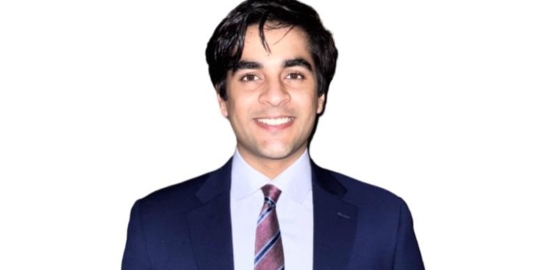 Udayveer Sekhon with dark hair, wearing a navy blue suit, light blue shirt, and striped tie, smiling against a plain white background.