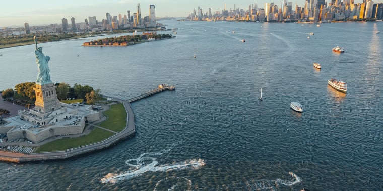 Aerial view of the Statue of Liberty on Liberty Island with boats in New York Harbor and the Manhattan skyline in the background under a clear sky.