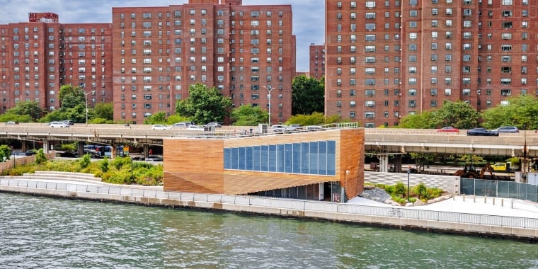 A modern wooden building for Solar One Environmental Education Center sits by the water with greenery around it, while tall red-brick apartment buildings and a highway with cars are visible in the background under a partly cloudy sky.