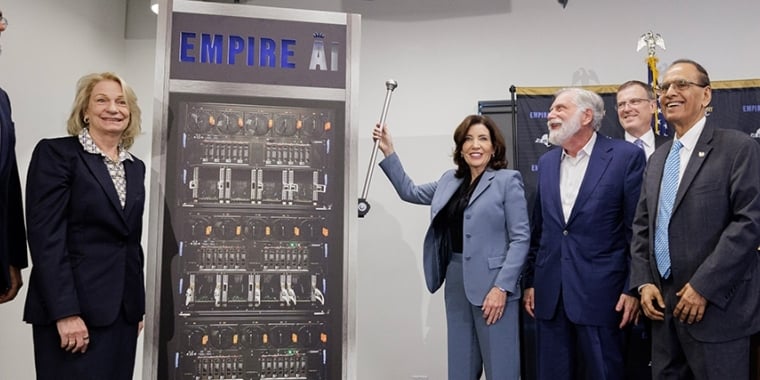 Six people in business attire stand smiling around a large sign labeled Empire AI, featuring an image of computer servers. One woman holds a gavel, suggesting an unveiling or announcement event.
