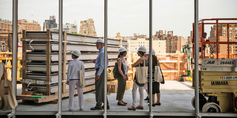 A group of people wearing hard hats stand inside a building under construction, looking out large windows with a city skyline in the background. Construction materials and equipment are visible around them.