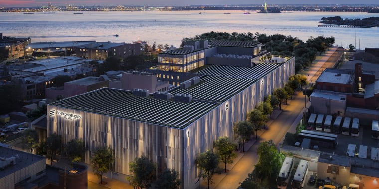 A modern building with a solar-paneled roof is surrounded by trees and city streets at sunset, overlooking a body of water with the Statue of Liberty visible in the distance.