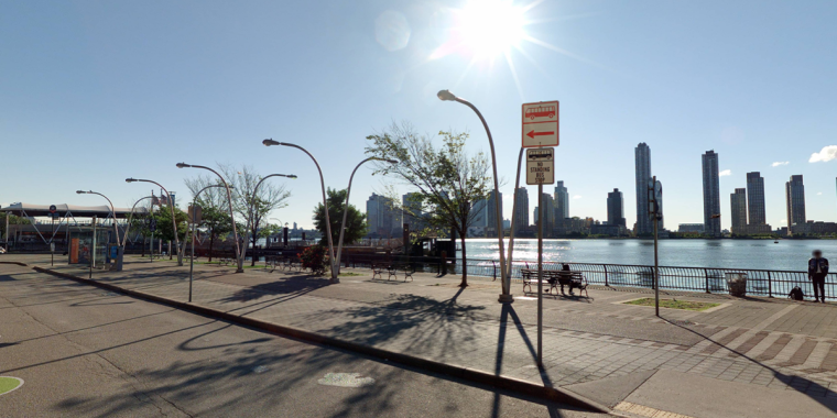 A sunny NYC scape featuring a river, tall buildings in the background, trees, street lamps, and empty benches along a riverside promenade with a few people scattered around.