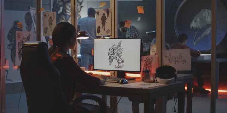 Woman sits at a desk, working on a digital drawing of a creature on a computer screen. The workspace is filled with sketches, a VR headset, and sticky notes. Other people work in the background behind glass walls.