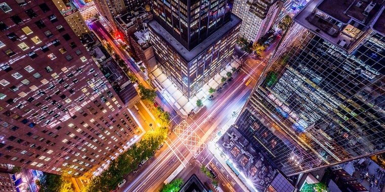 A vibrant, aerial night view of a New York City intersection surrounded by tall skyscrapers, with colorful lights from buildings and streaks of headlights and taillights showing busy street traffic below.