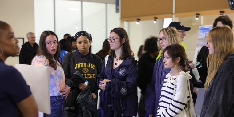 A group of young women stand together, listening to a woman at the Maritime Career Fair. Some wear jackets and casual clothes, and one has a “John Jay College” shirt. They appear attentive and engaged with the speaker.