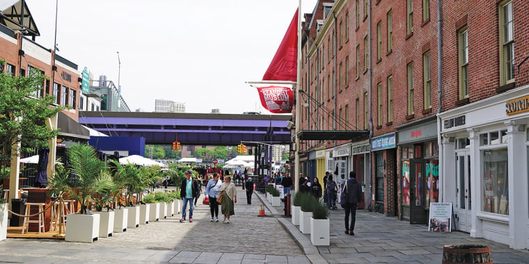 A cobblestone street with people walking, lined with red brick buildings and storefronts on the right, outdoor seating and plants on the left, and a red flag hanging above the sidewalk.