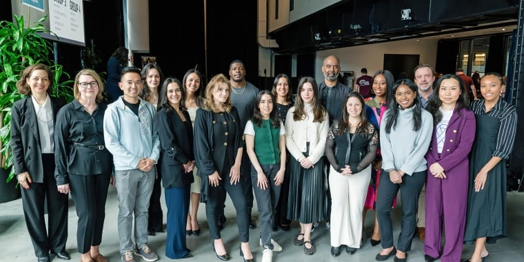 A diverse group of 18 people, both men and women, stand together indoors, posing and smiling for a group photo. They are dressed in business and business-casual attire.