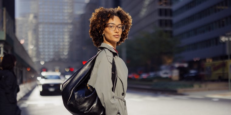 A person with curly hair, glasses, and a gray trench coat stands on a city street, looking over their shoulder. They have a large black bag and are surrounded by tall buildings with sunlight in the background.