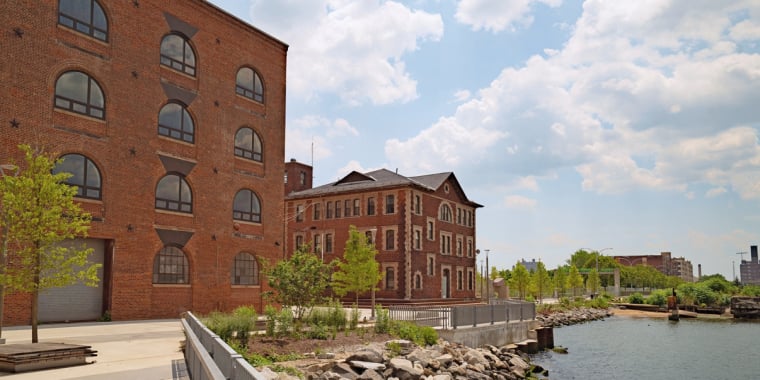 Red brick industrial buildings sit beside a river, surrounded by trees and rocks along the water’s edge. The sky is partly cloudy, and the area appears calm and well-maintained.