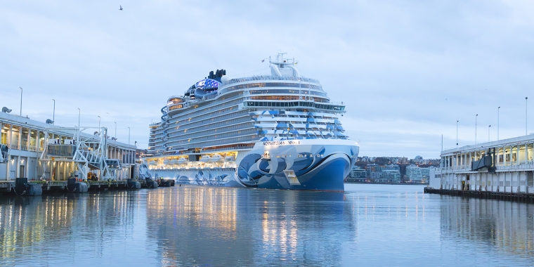 A large cruise ship, the Norwegian Prima, between two piers at the Manhattan Cruise Terminal on a calm, cloudy day. The ship features blue and white designs on its bow and is illuminated with lights. Buildings are visible in the background.