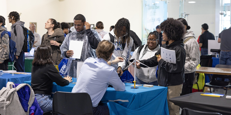 A group of students stands at a table interacting with seated staff at an indoor event. Papers and bags are visible, and other people are seen engaging in the background. The atmosphere appears lively and social.