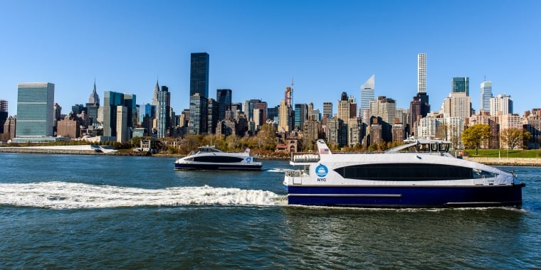Two NYC ferries travel on the East River with the Manhattan skyline, including tall skyscrapers and clear blue sky, in the background.