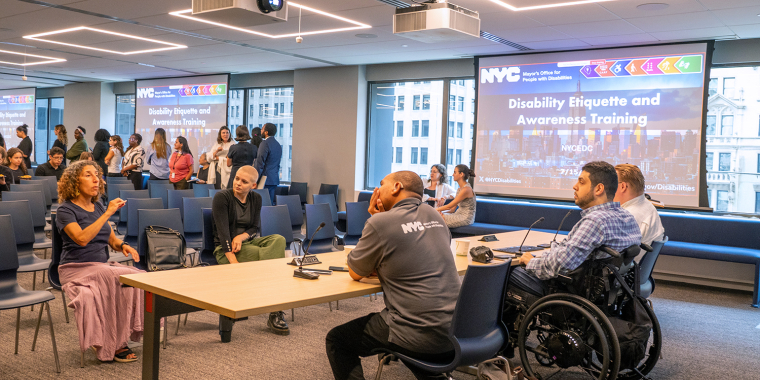 A diverse group of people, including individuals in wheelchairs, converse in a conference room before a presentation titled “Disability Etiquette and Awareness Training” on a projector screen.