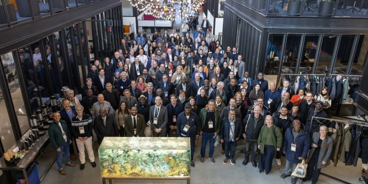 A large group of people posing for a photo inside a modern, industrial-style building with plants, glass offices, and a chandelier overhead. A large display case with natural materials is in the foreground.