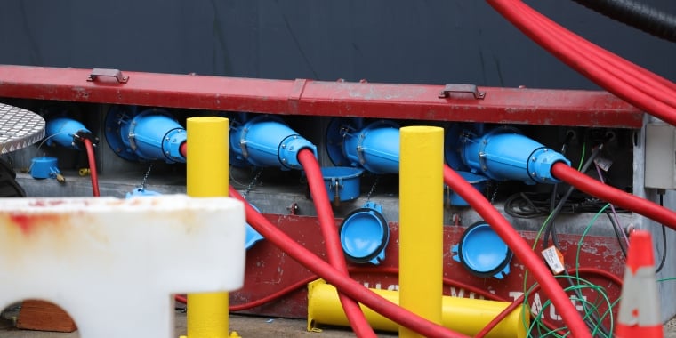 Multiple thick red cables are connected to blue industrial connectors mounted on a metal panel. Yellow protective posts are in front, and green wires and an orange safety cone are nearby. The background is a dark wall.