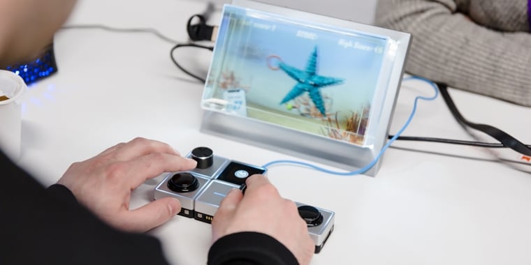 A person uses an adaptive game controller, playing a video game with a blue starfish on screen. Another person sits nearby at a white table, with cables and a drink visible.