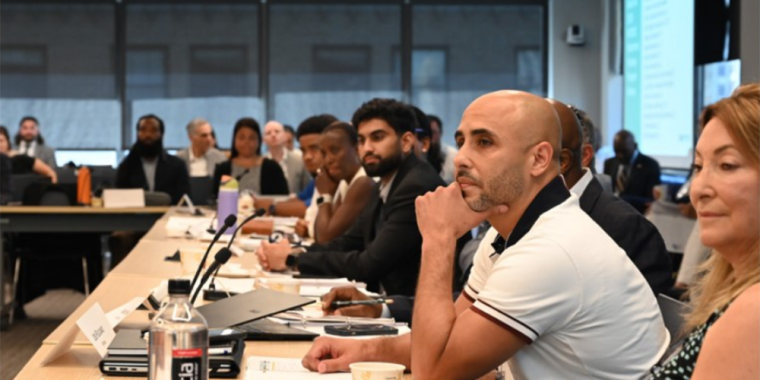 A diverse group of people sit attentively around a large conference table in a modern meeting room, listening to a presentation. Notebooks, cups, and water bottles are visible on the table.