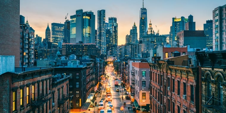 A New York City street lined with brick buildings stretches toward a skyline of tall, modern skyscrapers at sunset. Cars drive along the lit street, while the sky glows with warm evening light.